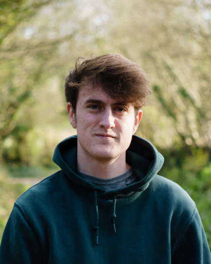 Person with brown hair that sweeps across his head to the right. Staring into the camera with deep green eyes and spots across his face. Wearing a dark green hoodie with a grey t-shirt underneath. Behind him the sun vibrantly illuminates the trees in a tunnel almost like vision either side of his head.